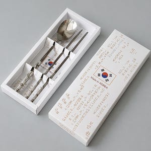 Könnte beinhalten: Ein Besteckset aus Edelstahl mit einem Löffel und einem Paar Essstäbchen, präsentiert in einer weißen rechteckigen Schachtel. Die Schachtel zeigt den Text Made in KOREA, eine südkoreanische Flagge und koreanische Schriftzeichen.