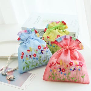 May include: Three drawstring pouches in light blue, yellow, and pink, each featuring a plaid pattern and embroidered colorful wildflowers. The bags are cinched at the top with matching fabric cords, showcasing detailed floral stitching.