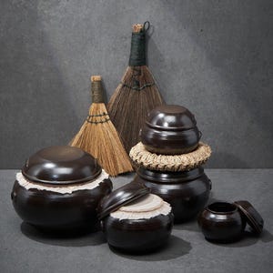 May include: A collection of dark brown glazed ceramic jars with matching lids, some featuring white fabric seals around the rims. Two natural fiber straw brooms with dark wrapped handles stand behind the jars, and one jar rests on a woven straw ring.