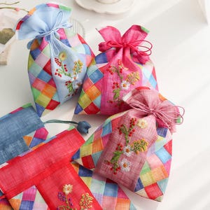 Puede incluir: Una colección de bolsas de tela con cordón que presentan un diseño de patchwork de cuadrados multicolores. Cada bolsa tiene un panel vertical de color sólido con intrincados bordados florales en verde, amarillo y blanco, cerradas con lazos.