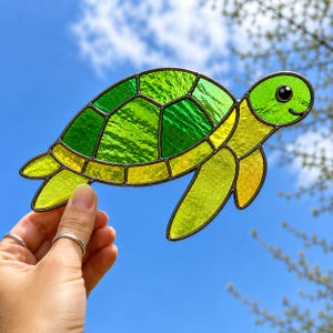 May include: A stained glass suncatcher shaped like a sea turtle with a textured green shell and yellow limbs. The piece is constructed with dark metal solder lines separating the individual glass segments, creating a clear outline of the turtle.