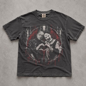 May include: A charcoal grey short-sleeve t-shirt featuring a graphic print of two horror characters forming a heart shape with their hands. The design is enclosed in a circular frame with red blood-like splatters and barbed wire accents.