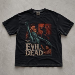 May include: A black short-sleeve t-shirt featuring a graphic print from the film The Evil Dead. The design includes a central teal figure with an outstretched hand, portraits of characters, and the text A FILM BY SAM RAIMI and THE EVIL DEAD.
