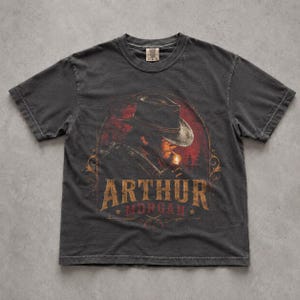 May include: A charcoal grey short-sleeve t-shirt featuring a graphic print of a person wearing a cowboy hat and holding a lit match. The text "ARTHUR MORGAN" is printed in a distressed gold font below the illustration, framed by decorative lines.