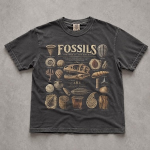 May include: A charcoal grey short-sleeve t-shirt featuring a beige graphic print of various fossils. The text reads FOSSILS HISTORY IN THE ROCKS. The design includes illustrations of a dinosaur skull, shells, leaves, and other prehistoric specimens.