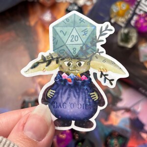 May include: A die-cut sticker featuring a small, green, goblin-like creature with large ears and a light blue twenty-sided die balanced on its head. The creature holds a dark purple pouch labeled BAG O' DICE filled with colorful miniature dice.
