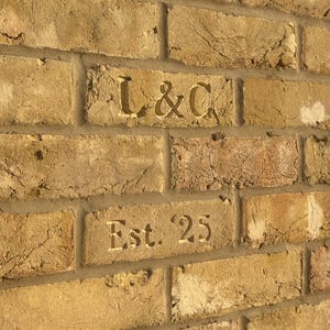 May include: A wall constructed of light brown, textured clay bricks. Two of the bricks feature engraved text. The top brick is engraved with the letters L & C, and the brick directly below it is engraved with the text Est. 25 in a serif font.