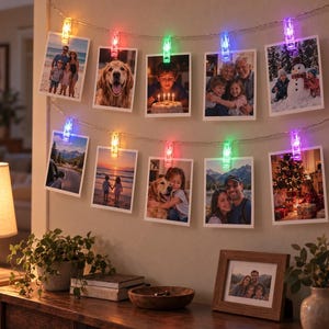 Puede incluir: Una cadena de luces LED con clips integrados que sostienen diez fotografías rectangulares. Los clips emiten varios colores, incluyendo rojo, verde, azul y morado, iluminando las fotos dispuestas en dos filas horizontales en la pared.