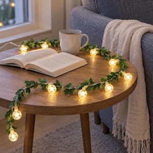 Op de afbeelding: Een snoer met LED-verlichting voorzien van kleine, doorzichtige bolvormige lampjes afgewisseld met groene kunstbladeren. Het lichtsnoer is in een cirkelvorm op een ronde houten tafel gelegd, rondom een open boek en een keramische mok.