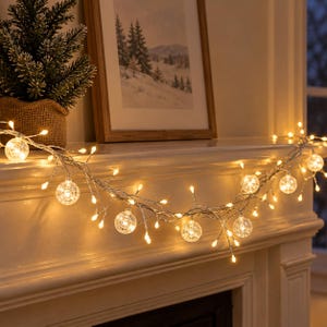 Peut inclure: Une guirlande lumineuse LED blanc chaud composée de globes en verre texturé transparent et de petites ampoules pointues sur un fil argenté, disposée horizontalement sur une cheminée pour créer un éclairage chaleureux.