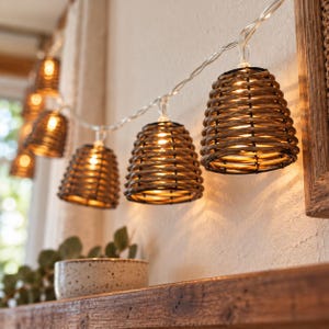 May include: A string of decorative lights featuring multiple bell-shaped shades crafted from woven brown rattan. Each shade houses a small, glowing bulb that emits a warm, soft light, connected by a thin, clear electrical cord.