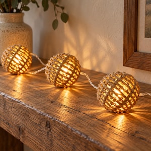 Peut inclure: Une guirlande lumineuse composée de trois sphères en corde de jute tressée de couleur beige. Chaque boule contient une ampoule diffusant une lumière chaude, reliées par un cordon électrique transparent posé sur une surface.