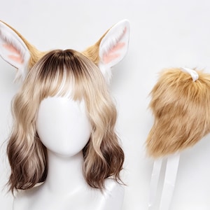 Puede incluir: Un conjunto que consta de un par de orejas de animal de piel sintética color canela y blanco unidas a una diadema, sobre una peluca rubia ondulada con raíces marrones, y una cola de piel sintética color canela con una cinta blanca.