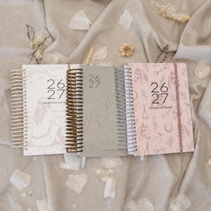May include: Three spiral-bound equestrian planners for the years 26-27. The left planner is white with a line drawing of a person, the center is grey with a faint illustration, and the right is pink with a repeating pattern of horses and gear.