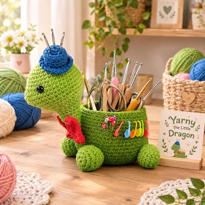 Puede incluir: Un organizador de ganchillo verde con forma de tortuga y un sombrero azul en la cabeza, que sostiene varios ganchillos de metal y tijeras. La tortuga tiene detalles rojos y marcadores de puntos de plástico de colores, con un letrero que dice Yarny the Little Dragon.