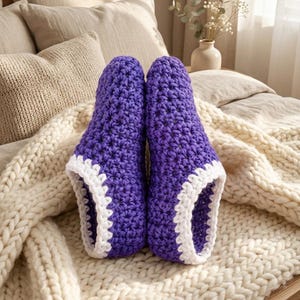 May include: A pair of purple crochet slippers featuring a textured star-like stitch pattern and a thick white crocheted trim around the ankle opening. The slippers are constructed from yarn and are positioned upright, showcasing the deep purple color and the contrasting white border.