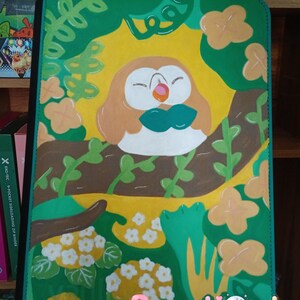 May include: A rectangular binder featuring a painted illustration of a round, tan and white bird character with a green bowtie resting on a brown tree branch. The background includes yellow sun rays, green foliage, and light orange flowers. Text reads Popp-Lionde.