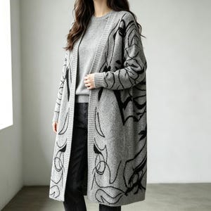 May include: A long, open-front knit cardigan in heather gray featuring a bold, black abstract line-art pattern throughout. The garment has long sleeves, a ribbed trim along the front opening, and a matching gray knit sweater worn underneath.