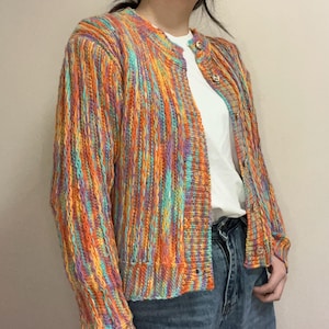 Può includere: Un cardigan in maglia a maniche lunghe caratterizzato da un vivace motivo a righe verticali multicolore nei toni dell'arancione, giallo, viola e turchese. Il capo presenta uno scollo rotondo, un'abbottonatura frontale e polsini a coste.