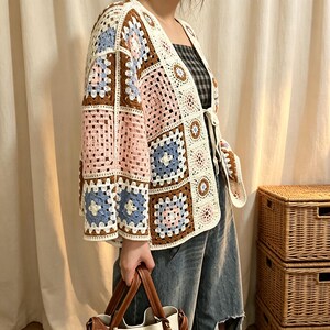 Può includere: Un cardigan all'uncinetto caratterizzato da un patchwork di piastrelle granny nei toni del marrone, azzurro, crema e rosa tenue. Il capo presenta un'apertura frontale con chiusura a laccio, maniche a tre quarti e un bordo color crema.