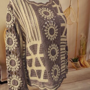 Può includere: Un maglione all'uncinetto a maniche lunghe con un design bicolore color malva e crema. Il capo presenta un motivo complesso di fiori circolari, strisce verticali e sezioni a griglia geometrica, realizzati con un intreccio di filati.