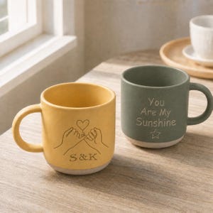 Peut inclure: Un mug en céramique jaune avec une illustration au trait de deux mains faisant une promesse avec un cœur et les lettres S&K, à côté d'un mug en céramique vert sauge avec le texte You Are My Sunshine et une icône d'étoile gravés sur le devant.