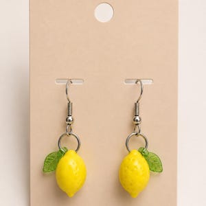 Puede incluir: Un par de pendientes colgantes con dijes de limón amarillo y pequeñas hojas verdes, unidos a ganchos de metal plateado. Los pendientes se presentan sobre una tarjeta rectangular de color beige con un orificio en la parte superior.