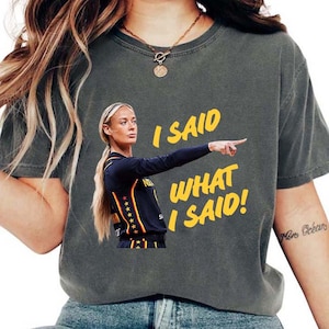 May include: A charcoal gray crew neck t-shirt featuring a graphic of a basketball player in a dark blue jersey with yellow and red accents, pointing to the right. The yellow text next to the player reads, I SAID WHAT I SAID!