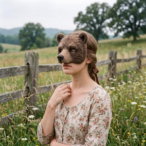 Puede incluir: Una máscara de oso pardo hecha de fieltro texturizado que cubre la parte superior del rostro, con orejas redondeadas, una nariz oscura y cintas marrones. La máscara se lleva con una camisa de manga larga clara con un pequeño estampado floral rojo y verde.