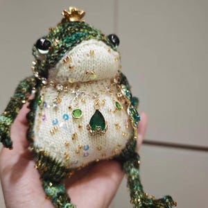 May include: A handcrafted knitted frog figurine featuring a cream-colored belly adorned with gold beads and sequins. The frog wears a delicate gold-toned necklace with small clear crystals and a prominent teardrop-shaped green pendant with a gold crown.