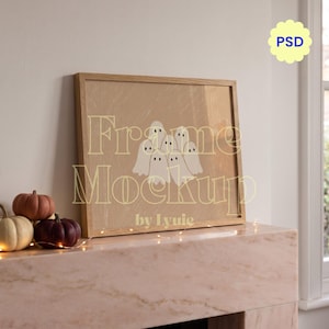 May include: A rectangular wooden picture frame containing a tan print with a group of white ghost illustrations. The frame is positioned on a light pink mantelpiece next to small pumpkins and string lights. Text on the image reads Frame Mockup by Lyuie.