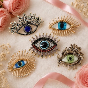 May include: Five eye-shaped brooches in various designs. One features a dark blue center with black crystals, another has a blue iris, one displays a black center with turquoise and red accents, one has a blue iris with rays, and one has a green iris.