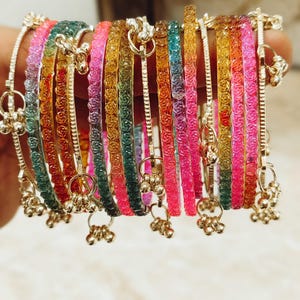 May include: A collection of multi-coloured bangles featuring a textured rose pattern in shades of pink, orange, yellow, green, and blue. Each bangle is accented with small, dangling metallic charms that produce a jingling sound when moved.