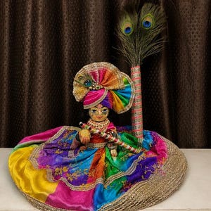 May include: A small figurine of a deity with a painted face, wearing a vibrant, multi-coloured silk outfit and a matching pleated turban. The figure holds a small flute and is positioned next to a tall, slender pole topped with peacock feathers.