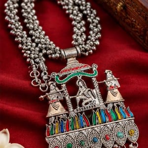 May include: A silver-toned necklace featuring a multi-strand beaded chain and a large, ornate pendant. The pendant depicts a central figure flanked by two musicians, accented with red, green, and blue enamel, and finished with dangling beads.
