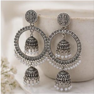 May include: A pair of silver-toned jhumka earrings featuring a circular frame with intricate embossed patterns. Each earring includes a central bell-shaped jhumka and a smaller dangling jhumka at the bottom, both adorned with tiny white beads.