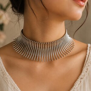 Könnte beinhalten: Eine silberfarbene Halskette, die aus einer Reihe vertikaler, stabförmiger Metallstangen besteht, die fächerförmig um den Hals angeordnet sind. Die einzelnen Stangen sind oben durch eine horizontale Kette verbunden, was ein strukturiertes, fransenartiges Aussehen erzeugt, das auf dem Schlüsselbeinbereich aufliegt.