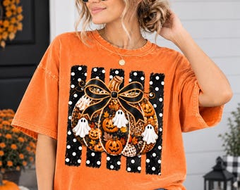 May include: An orange short-sleeve t-shirt featuring a graphic print of a large pumpkin decorated with a black bow, small white ghosts, and autumn leaves. The design is set against a background of vertical black stripes with white polka dots.
