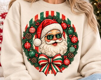 May include: A beige crewneck sweatshirt featuring a graphic of Santa Claus wearing sunglasses with tree reflections. Santa is framed by a Christmas wreath decorated with red and green ornaments, pinecones, and a large striped bow at the bottom.