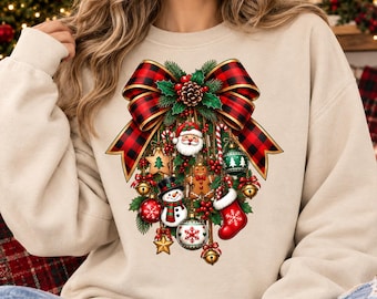 May include: A beige crewneck sweatshirt featuring a large graphic print of a red and black buffalo plaid bow at the top, with hanging Christmas ornaments including a Santa face, a gingerbread man, a snowman, a stocking, and gold bells.