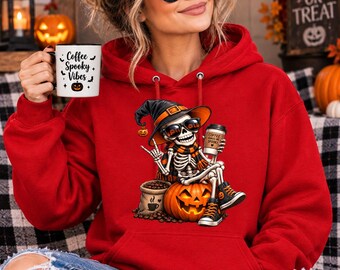 May include: A bright red hooded sweatshirt featuring a graphic of a skeleton wearing a witch hat, sunglasses, and a scarf, sitting on a pumpkin while holding a coffee cup. The white mug held in the hand displays the text Coffee Spooky Vibes.