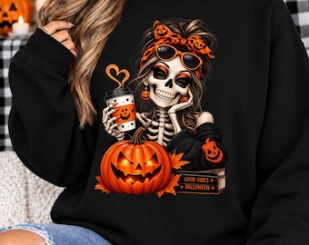 May include: A black sweatshirt featuring a graphic of a skeleton with a pumpkin-patterned headband and sunglasses, holding a coffee cup with a heart-shaped steam. A glowing jack-o'-lantern sits below, with books labeled Good Vibes Halloween.