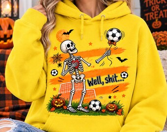May include: A bright yellow hooded sweatshirt featuring a graphic of a skeleton holding a soccer ball and a red heart. The design includes pumpkins, a spider web, bats, and the text "Well, shit..." printed in black over orange brush strokes.