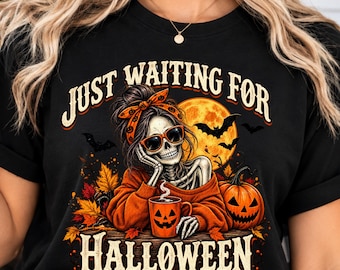 May include: A black t-shirt featuring a graphic of a skeleton wearing sunglasses and an orange headband, resting its chin on a hand. The skeleton holds an orange jack-o'-lantern mug. Text reads JUST WAITING FOR HALLOWEEN with a full moon and bats.