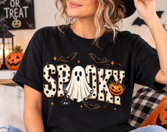 May include: A black t-shirt featuring the word SPOOKY in large white letters with black polka dots. The design includes a central white ghost, a bright orange jack-o'-lantern, several black bats, small orange stars, and a spider hanging from a web.