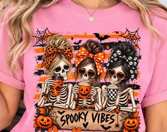 May include: A light pink t-shirt featuring a graphic of three skeletons with messy buns and large bows, wearing heart-shaped sunglasses and holding coffee cups. The design includes orange and black stripes, pumpkins, and the text SPOOKY VIBES.