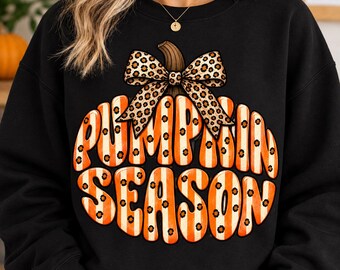 May include: A black long-sleeve sweatshirt featuring a large graphic of a pumpkin with orange and white vertical stripes, a brown stem, and a leopard print bow. The text PUMPKIN SEASON is printed in a bold, rounded font across the pumpkin graphic.