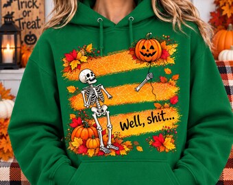 May include: A green hooded sweatshirt featuring a graphic print of a skeleton holding a string attached to a jack-o'-lantern. The design includes orange stripes, autumn leaves, and the text "Well, shit..." printed in black script lettering.