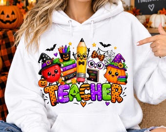 May include: A white hooded sweatshirt featuring a colorful graphic of a smiling apple, a pencil, a ghost, and a pumpkin wearing hats. The design includes a stack of books labeled Teach, Inspire, Change, Lives and the word TEACHER in large letters.