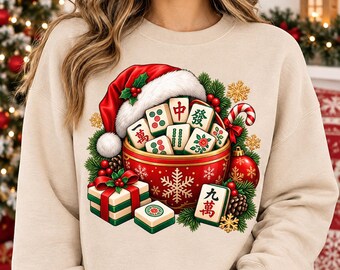 May include: A beige crewneck sweatshirt featuring a graphic of a red bowl filled with Mahjong tiles, topped with a Santa hat. The design includes green pine branches, a candy cane, a red gift box with a green ribbon, and gold snowflake accents.